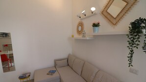 A living room with a beige sofa, a small table with a magazine, a red chair, a wall-mounted shelf with decorative items, and a hanging plant. - Charming Holiday Apartment "Casa Vacanze Aura" with Air Conditioning (Otranto)
