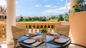 A round table with chairs, a potted plant, and breakfast items on a balcony overlooking a cityscape.