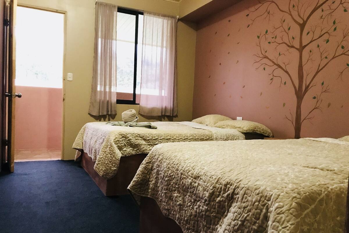 A room with two beds, a window with curtains, a door, and a wall with a tree mural.