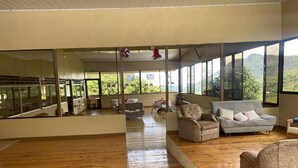 Una habitación amplia con suelo de madera, un sofá gris, un sillón color beige y un sofá blanco con cojines. Grandes ventanales ofrecen vistas a zonas verdes y colinas.
