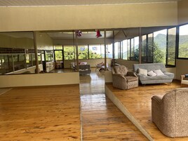 A spacious room with wooden flooring, a grey sofa, a beige armchair, and a white sofa with cushions. Large windows offer a view of greenery and hills.