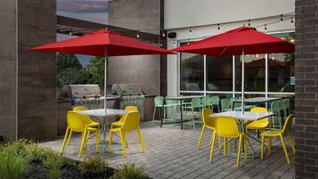 An outdoor patio with yellow chairs and red umbrellas, a grill, and a brick wall.