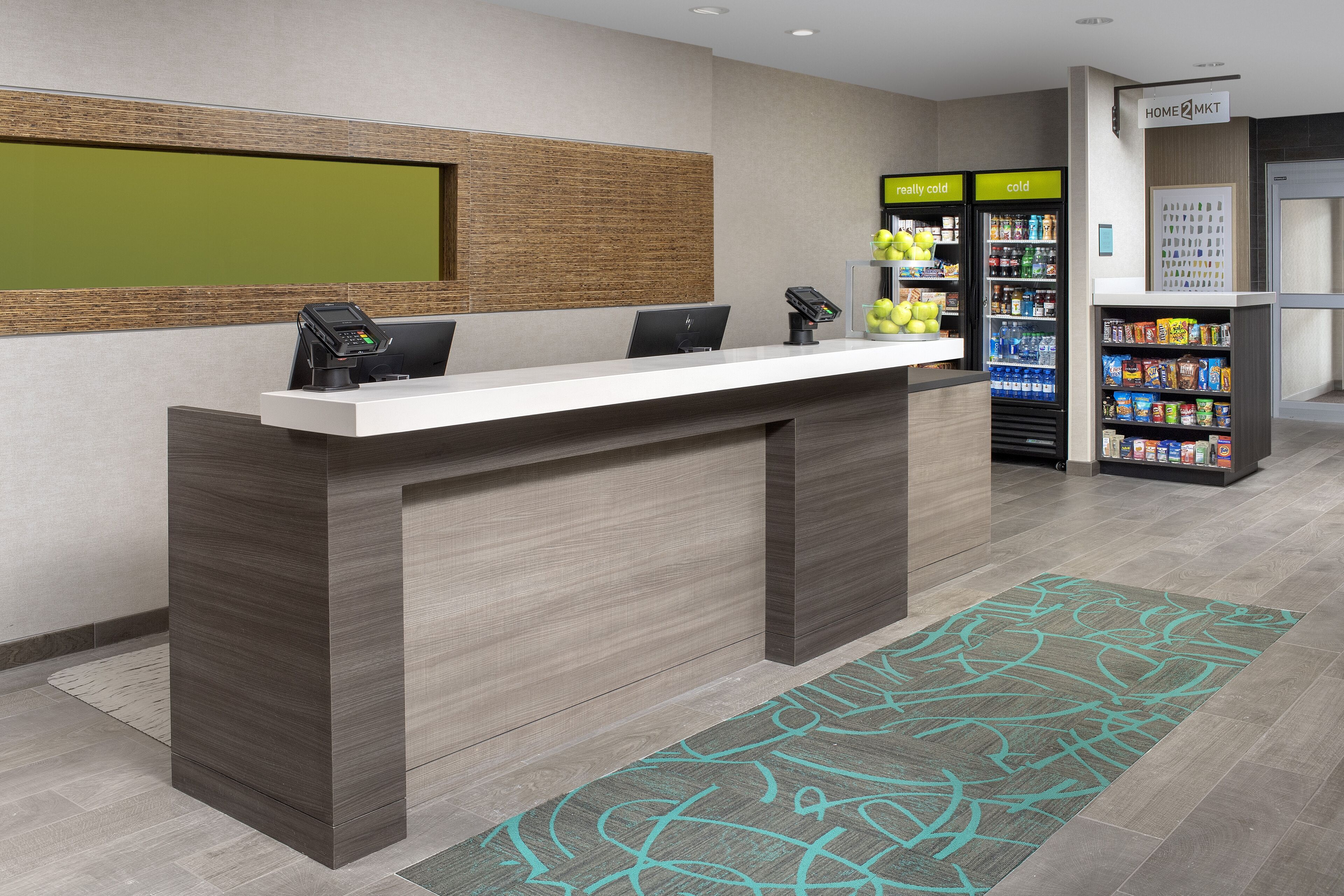 Un mostrador de recepción con encimera blanca, un terminal de pago digital y un pequeño espacio de descanso. Se ven máquinas expendedoras y un refrigerador al fondo.