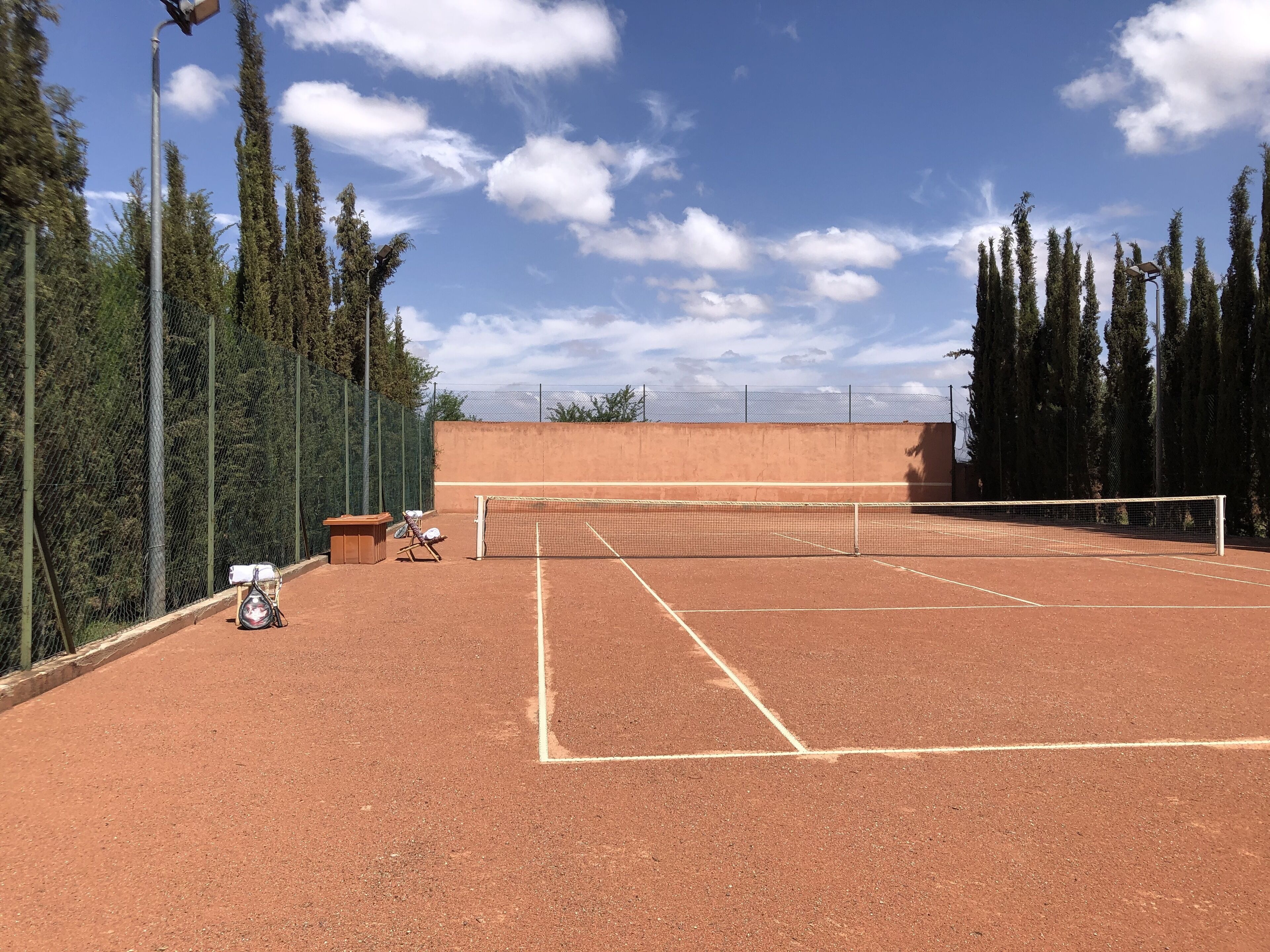 An outdoor tennis court with a net, surrounded by tall trees and a fence.