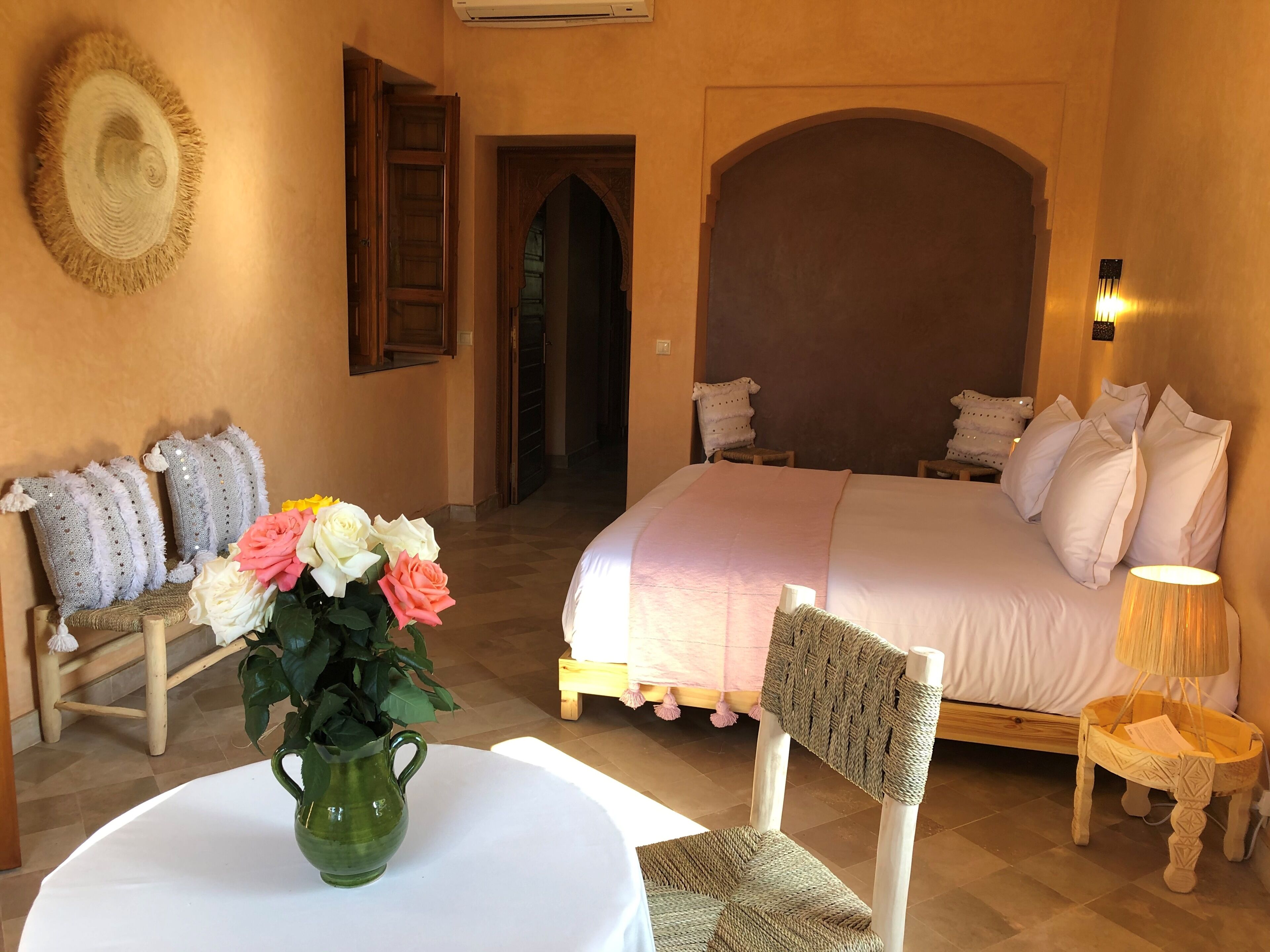 A bedroom with a large bed, a wooden chair, a small table with a vase of flowers, and a wall-mounted lamp.