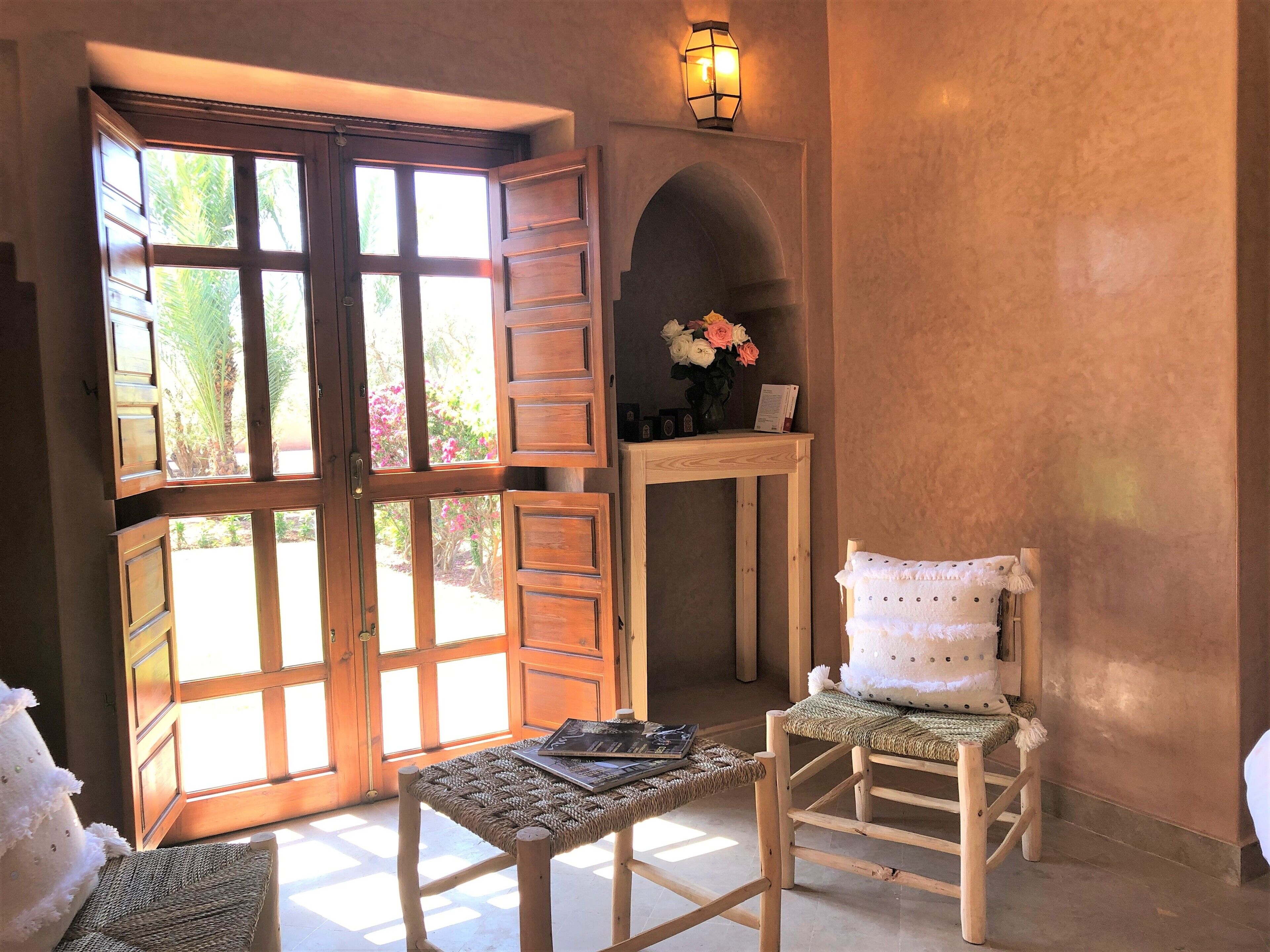 A room with a large wooden door open to a garden, a small table with a woven chair, and a wall-mounted light fixture.