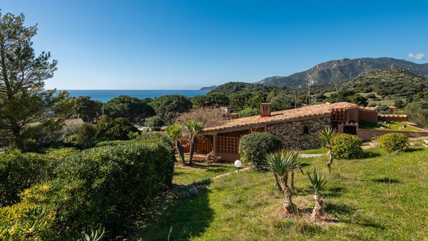 A hillside villa with stone walls, a garden, and a view of the sea.