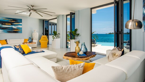 Una sala de estar moderna con un sofá blanco, cojines amarillos, una mesa de centro de madera con objetos decorativos y vistas al océano a través de grandes puertas de vidrio.
