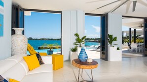 Una sala de estar moderna con un sofá blanco, cojines amarillos y un jarrón grande. Se ve una piscina y una playa a través de amplias ventanas.