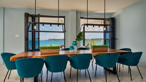 Un comedor con mesa de madera y sillas color turquesa, ventanas grandes con marcos negros, y lámparas colgantes.