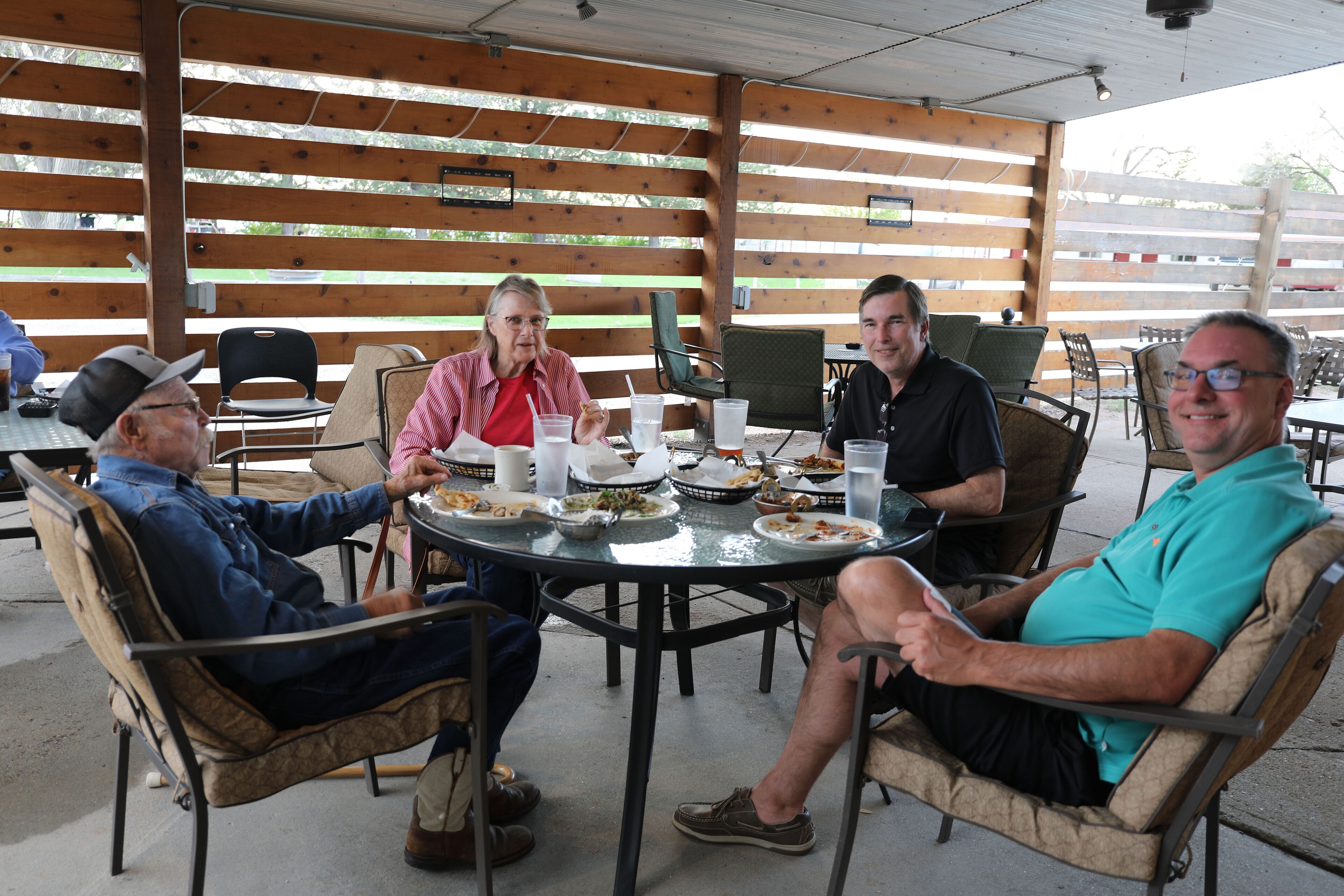 A group of people seated at a round table with plates of food, in a covered outdoor area with wooden slats.
