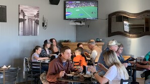 A restaurant interior with patrons seated at tables, a television mounted on the wall displaying a sports game, and a framed picture of a cityscape on the wall. - Raine Motel (Valentine)
