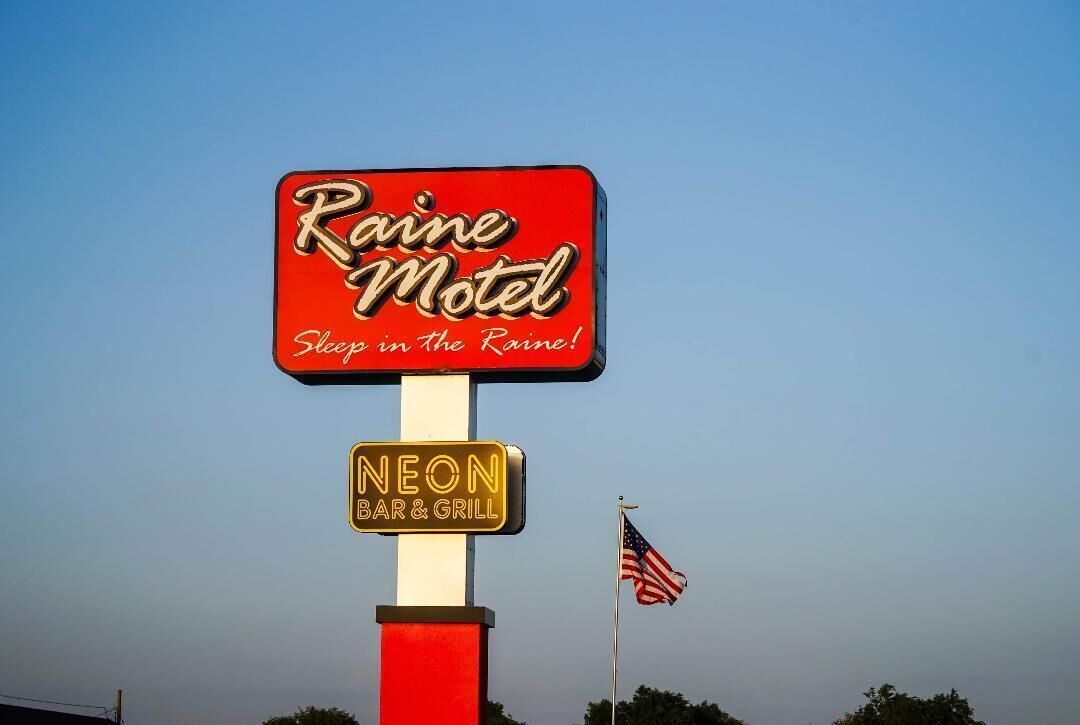 Et motel skilt med teksten "Raine Motel" og "Sleep in the Raine!". Et neonskilt for en bar og grill, samt et amerikansk flag.