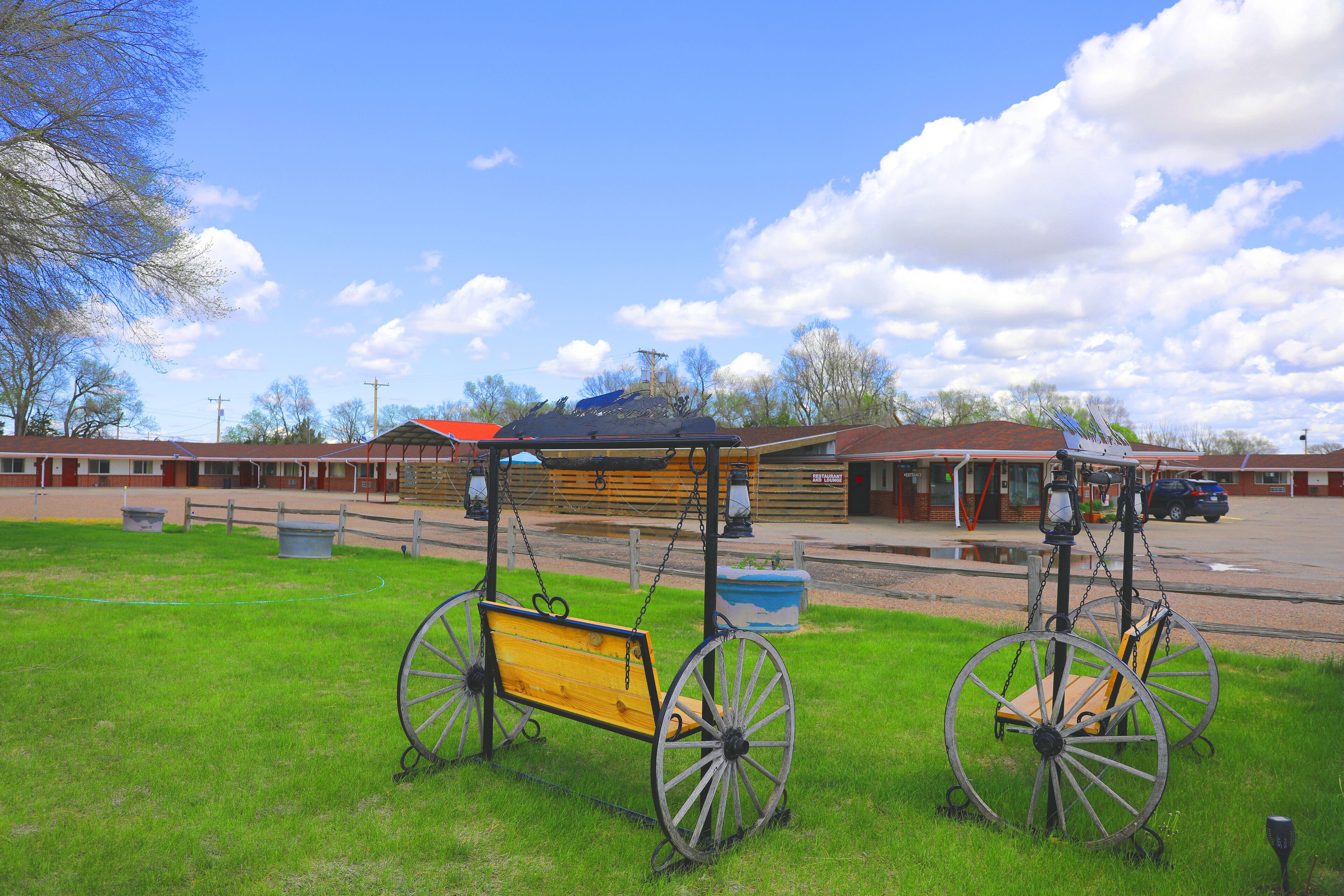 A rustic wooden bench with a metal frame and wheels, situated on a grassy area with a building in the background.