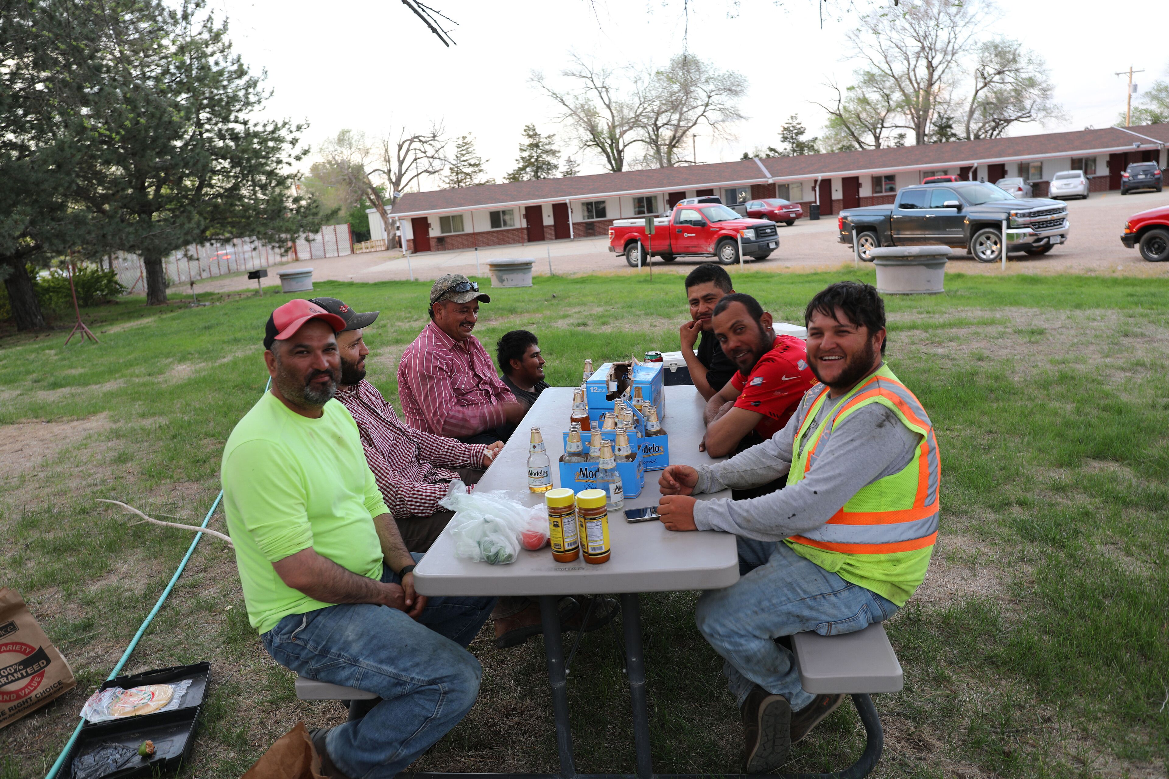 A group of people seated around a table with food and drinks, outdoors with a building and vehicles in the background.