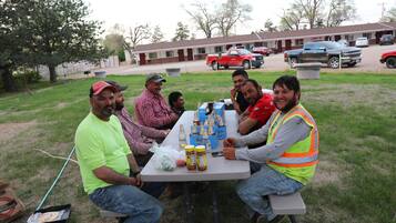 A group of people seated around a table with food and drinks, outdoors with a building and vehicles in the background.