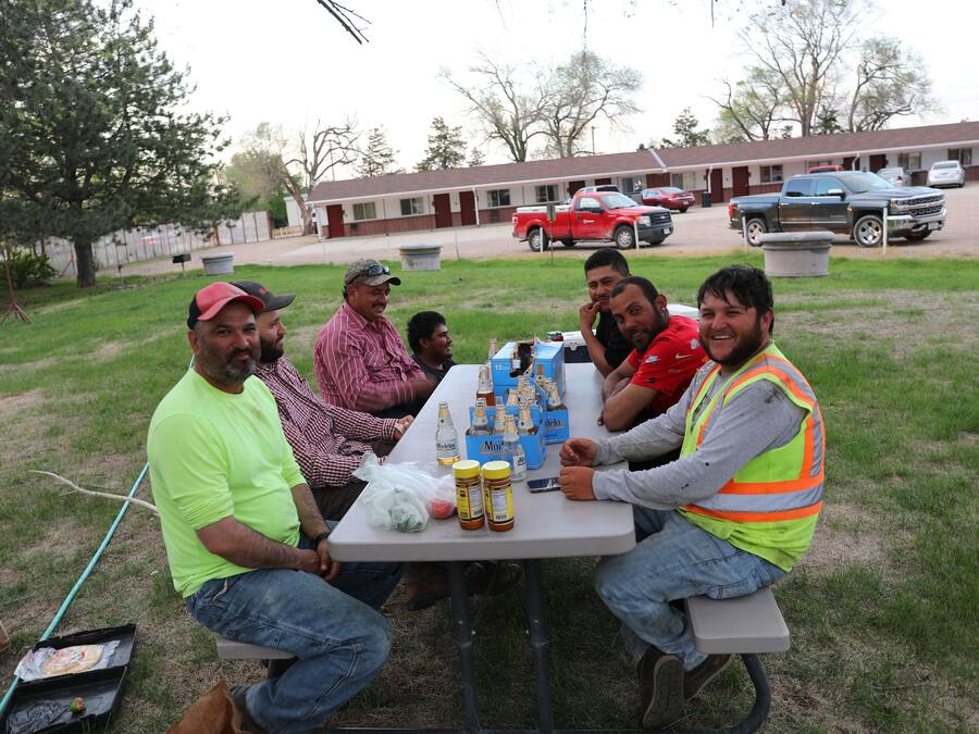 A group of people seated around a table with food and drinks, outdoors with a building and vehicles in the background.
