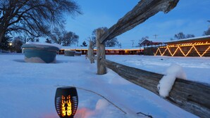 A snow-covered outdoor area with a wooden fence, a bench, and a building with illuminated lights in the background. - Raine Motel (Valentine)