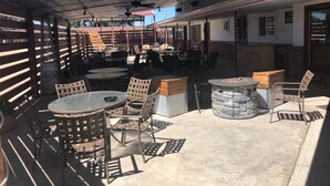 An outdoor patio area with round tables and chairs, a stone fire pit, and a covered seating area with a metal roof. - Raine Motel (Valentine)