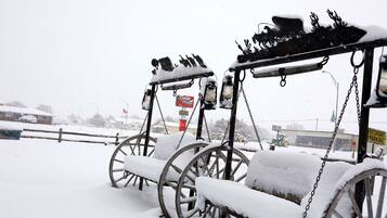 Zona exterior con nieve, con sillas y mesas de metal, un letrero que indica un estacionamiento y una vista distante de un edificio y vehículos.