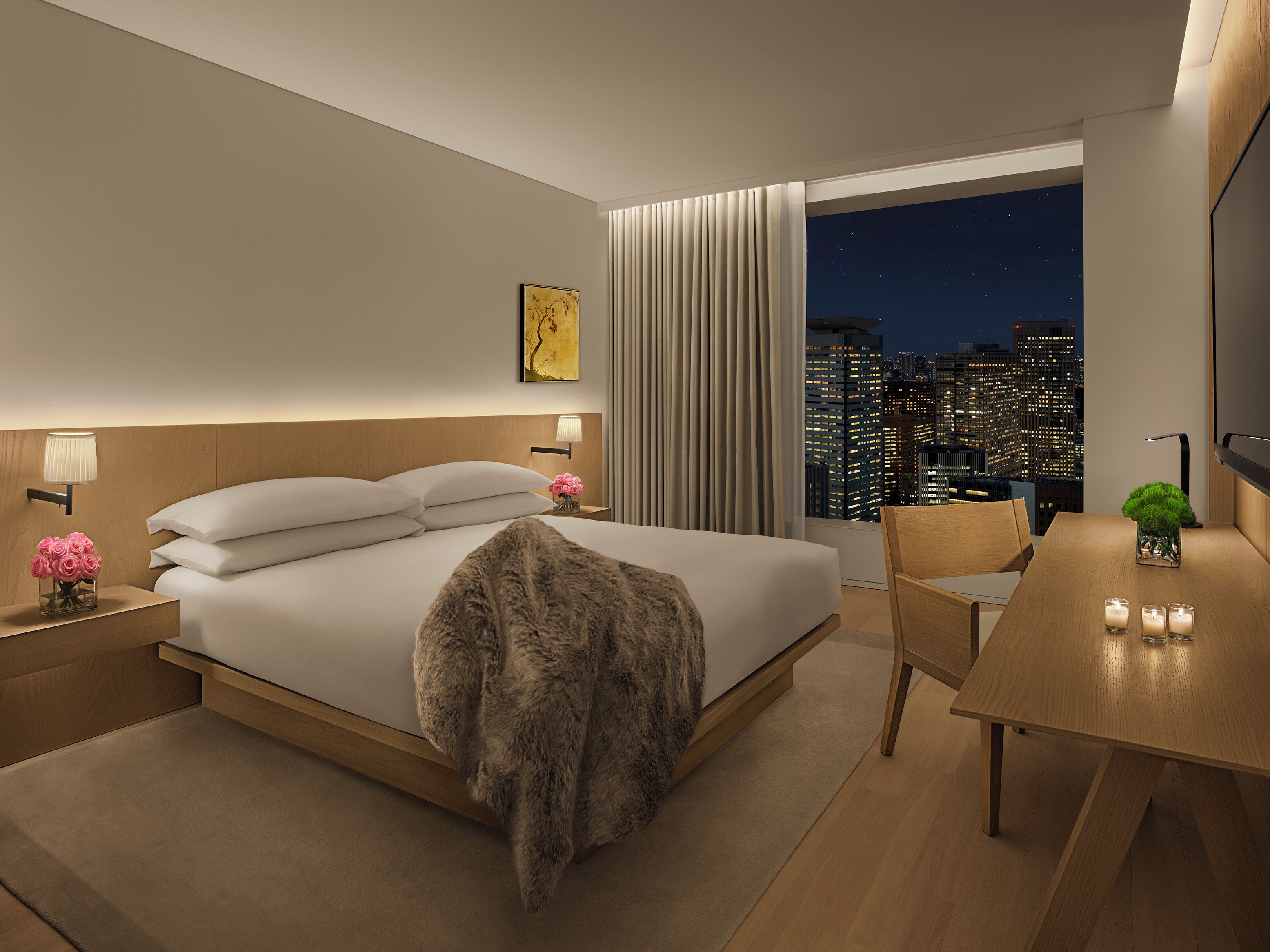 A modern hotel room with a large bed, a desk, a view of the city at night, and a painting on the wall.