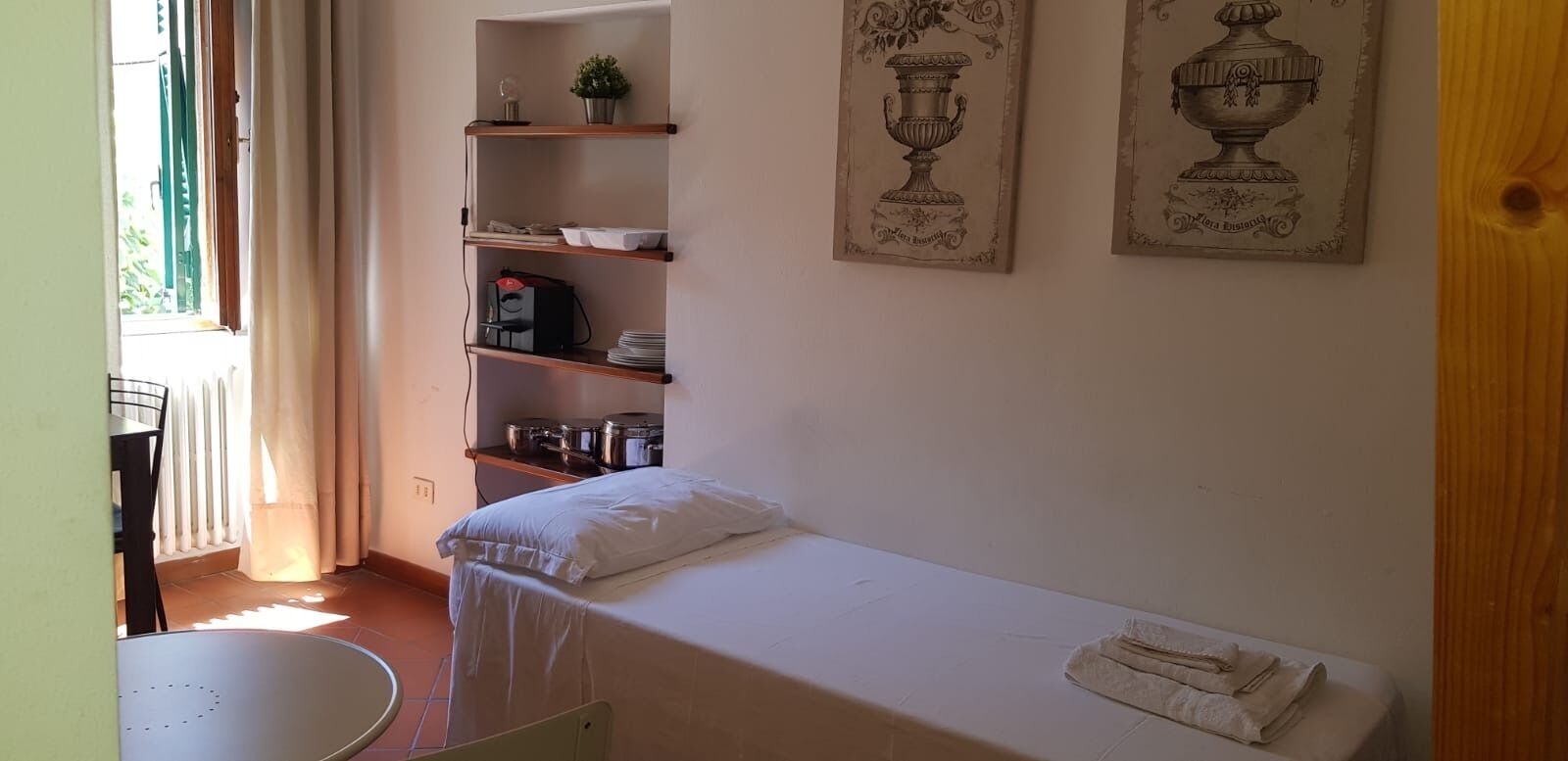 A small, tidy room with a single bed, a table, and a shelf with kitchenware. - Nido del Corso apartment (Pontedera)