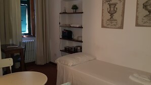 A small, simple room with a single bed, a chair, a table, and a shelf with kitchen items. - Nido del Corso apartment (Pontedera)