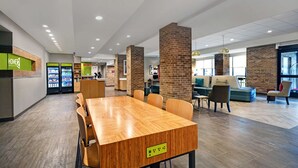 A modern indoor space with a wooden conference table, seating, a refreshment area, and brick pillars. - Home2 Suites by Hilton Liberty NE Kansas City (Liberty)
