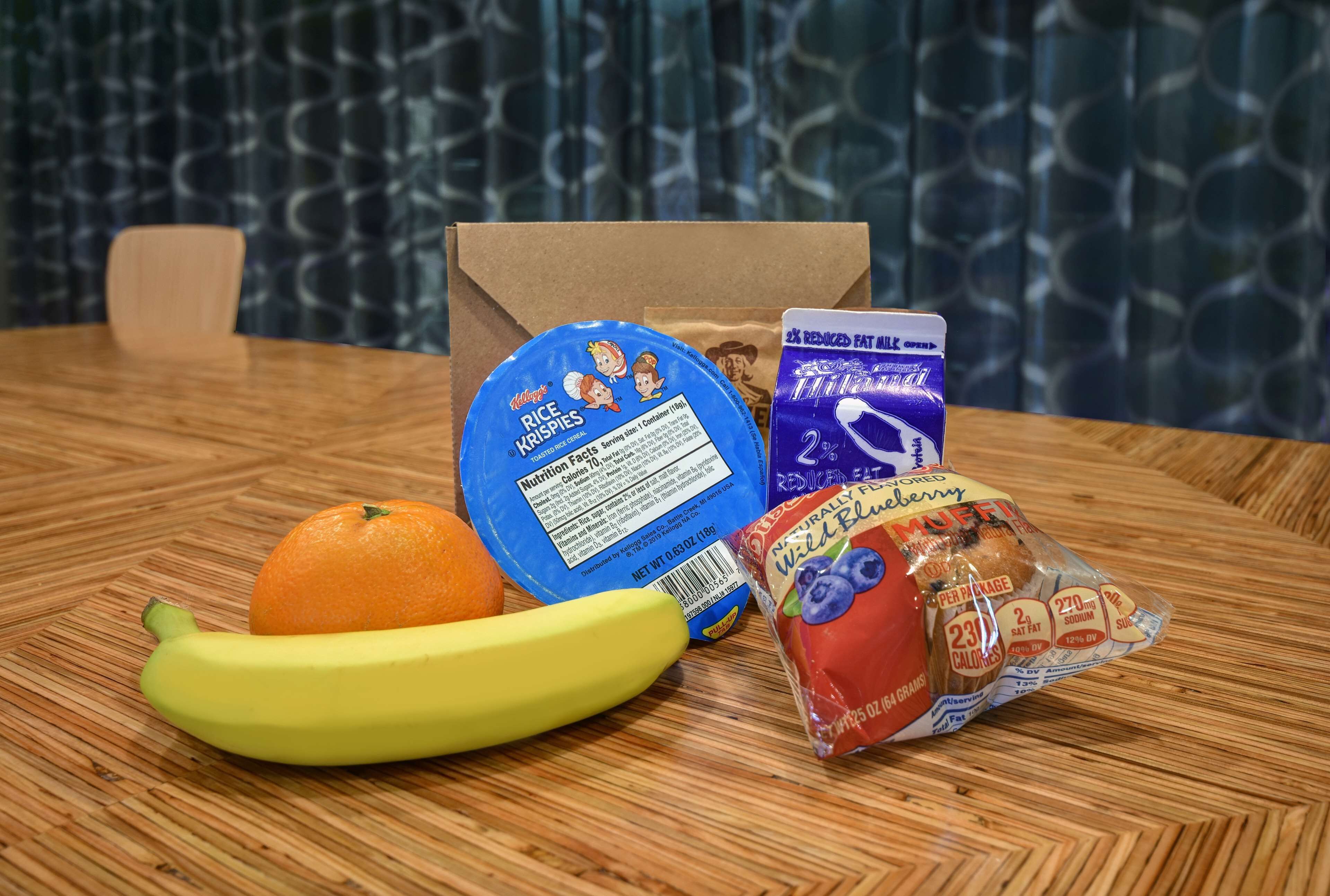 A table with a banana, an orange, a blue container of rice krispies cereal, a package of blueberry muffins, and a purple box of reduced-fat milk.