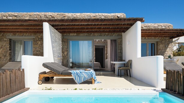 A poolside area with a lounge chair, a table, and a view of a stone building. - Aegean Hospitality Mykonos (Mykonos)