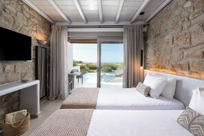 A bedroom with two beds, a TV, a stone wall, and a view of the outdoors. - Aegean Hospitality Mykonos (Mykonos)