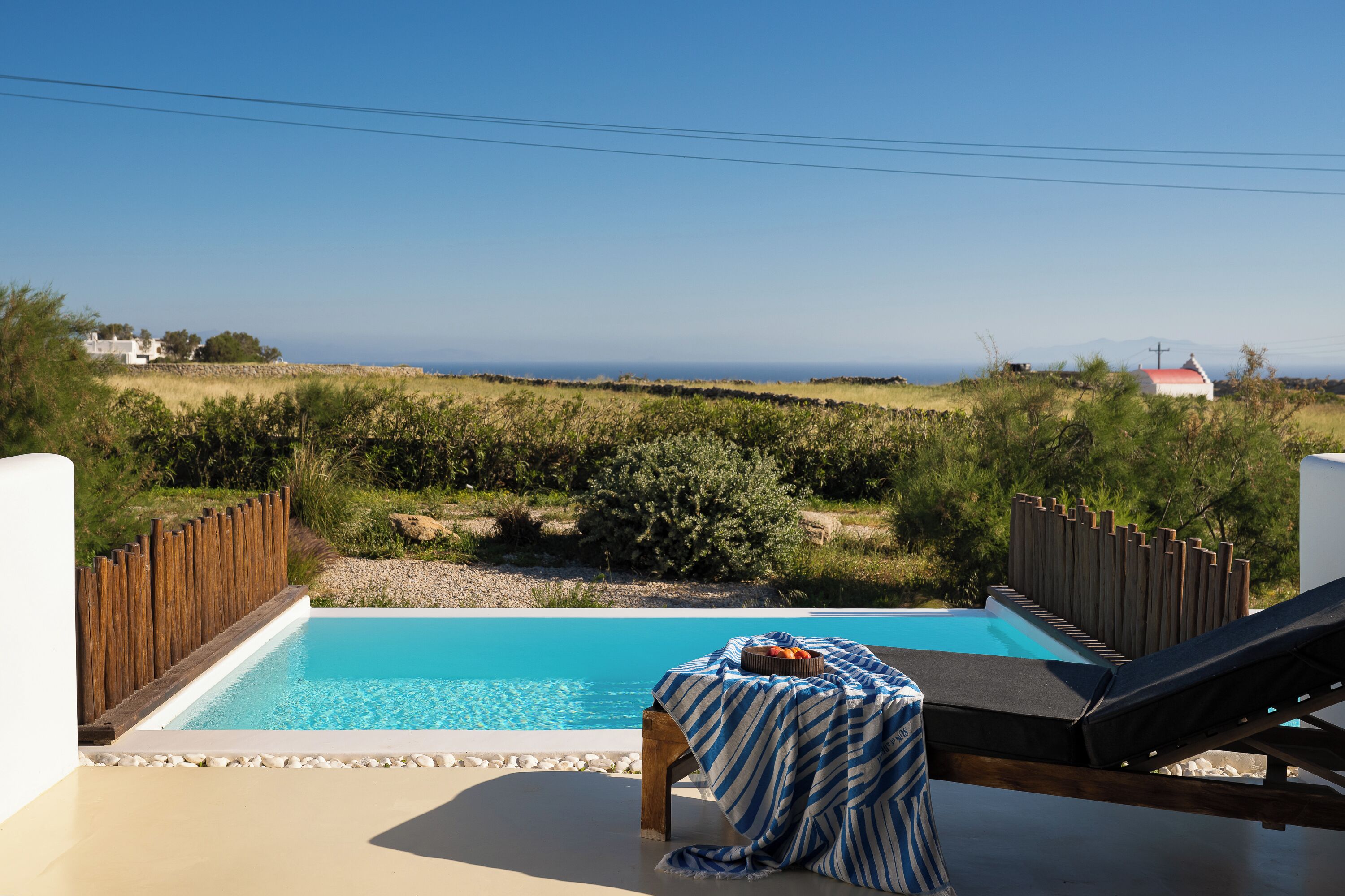 A poolside area with a lounge chair, a table with a towel, and a view of the sea and distant buildings.