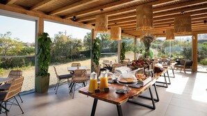 A dining area with a long wooden table, wicker chairs, and a view of the outdoors. - Aegean Hospitality Mykonos (Mykonos)