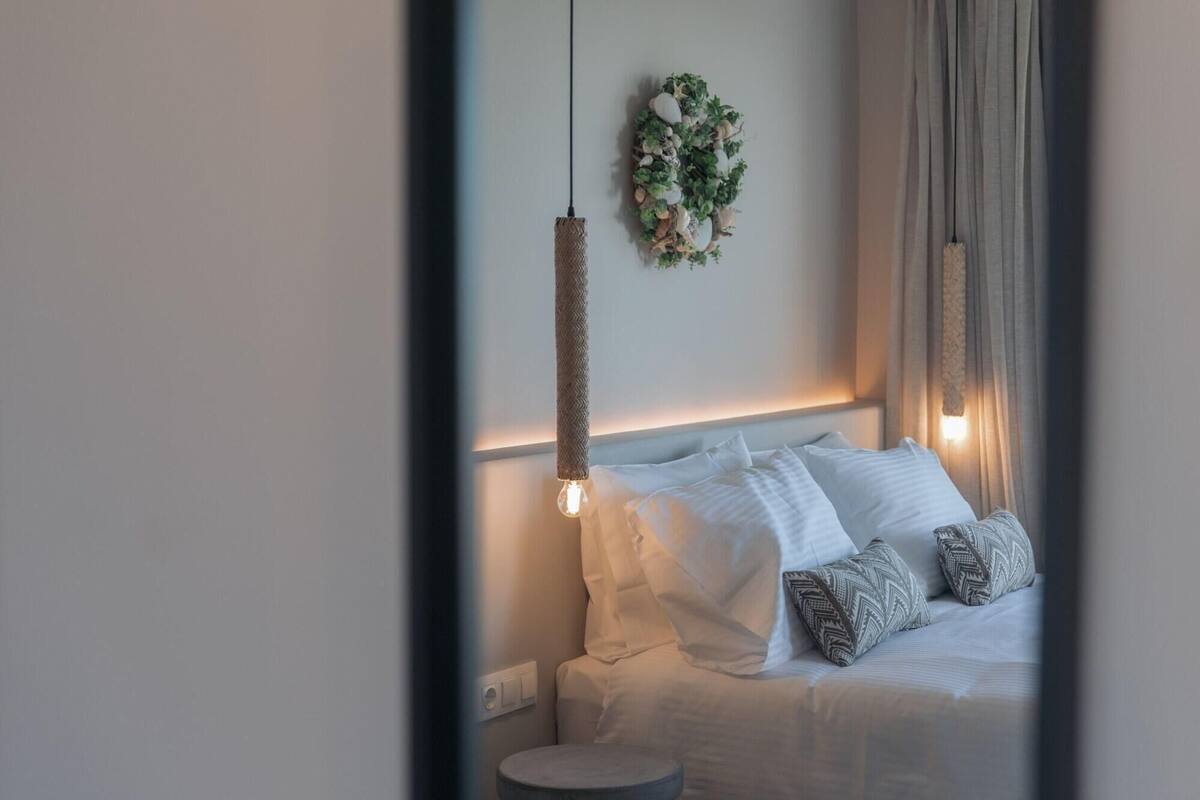 Ein Schlafzimmer mit einem Bett, Kissen, einer Pendelleuchte, einem kleinen runden Tisch und einer Wandpflanze.