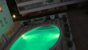 An outdoor swimming pool with a green-lit interior, surrounded by a tiled deck and a low wall with plants. - Loc Apartment Ben T3 - 6 to 8 people in La Grande Motte (LA GRANDE MOTTE)