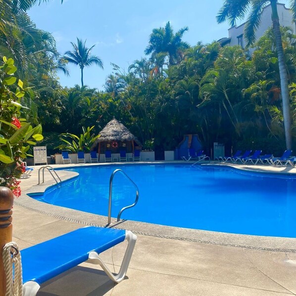 A swimming pool with lounge chairs and a thatched-roof hut, surrounded by palm trees and tropical vegetation.