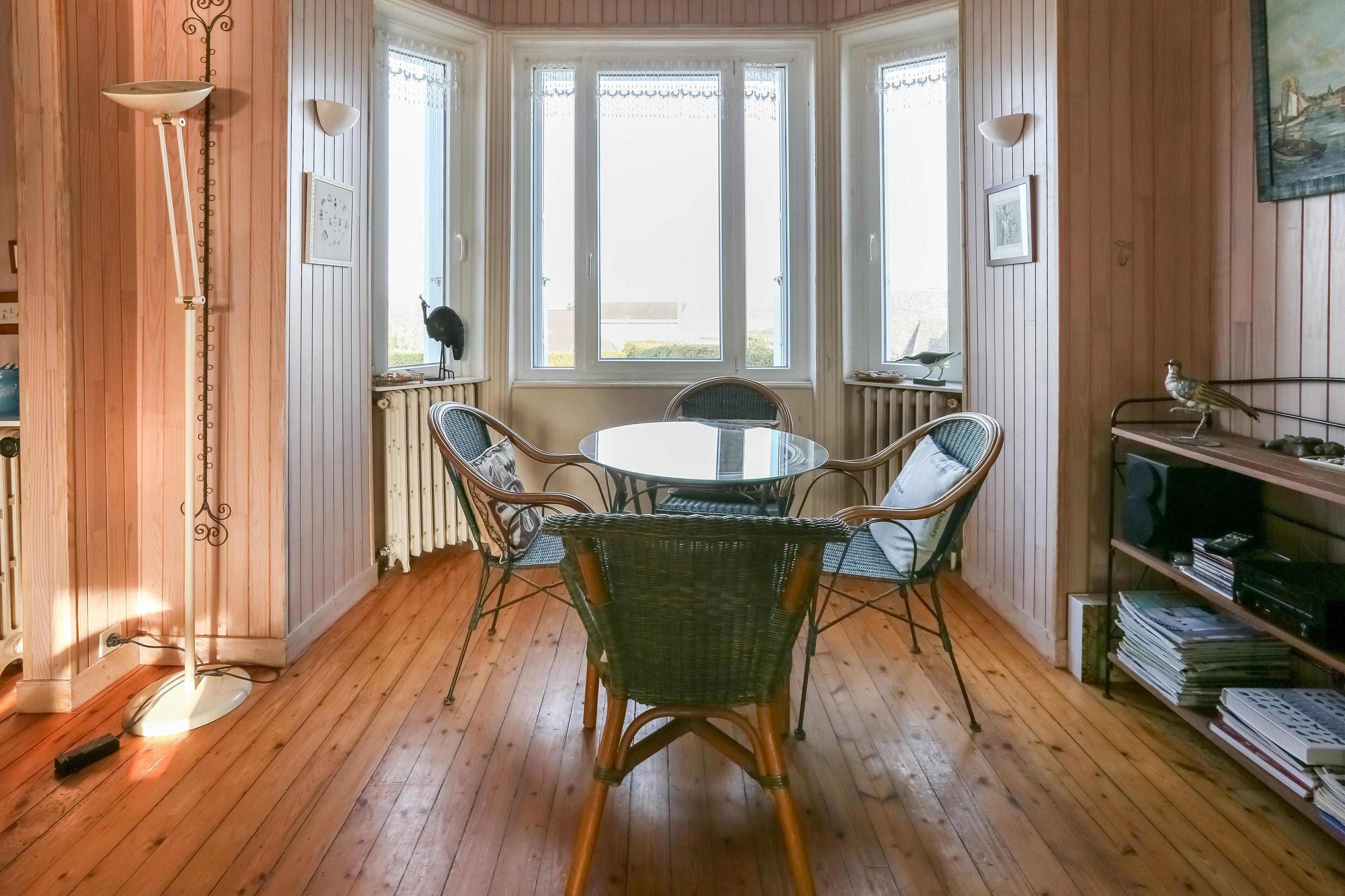 A room with a round table surrounded by wicker chairs, a radiator, and a bookshelf with books.