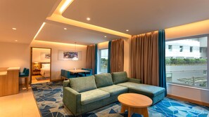 A modern hotel room with a sofa, a round wooden table, a dining area, and a large window with curtains.