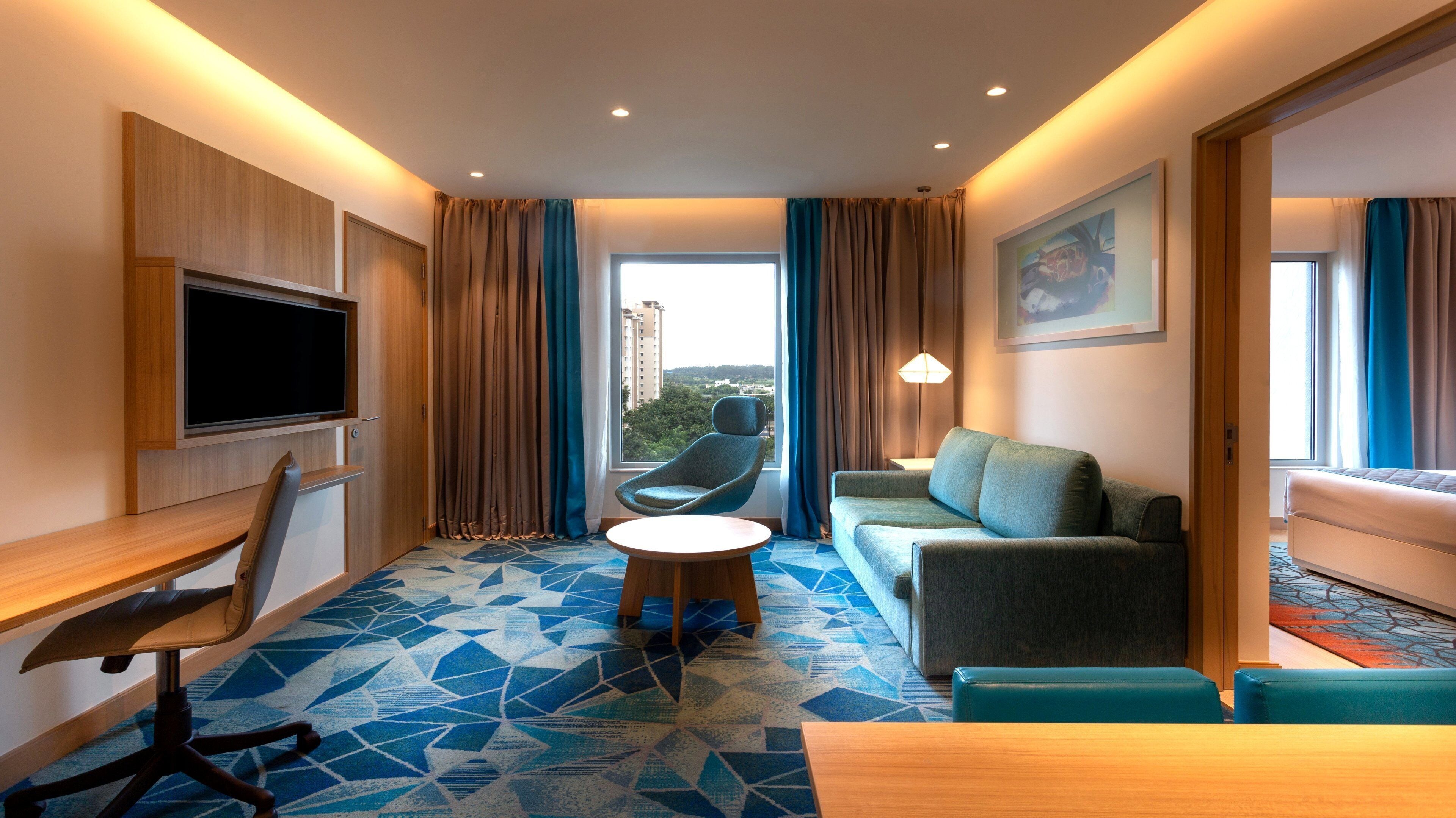 A hotel room with a TV, a sofa, a chair, a small table, and a view of buildings through the window.