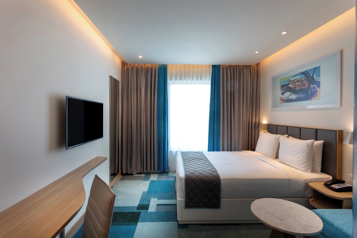 Een hotelkamer met een bed, nachtkastje, stoel, televisie en een schilderij aan de muur.