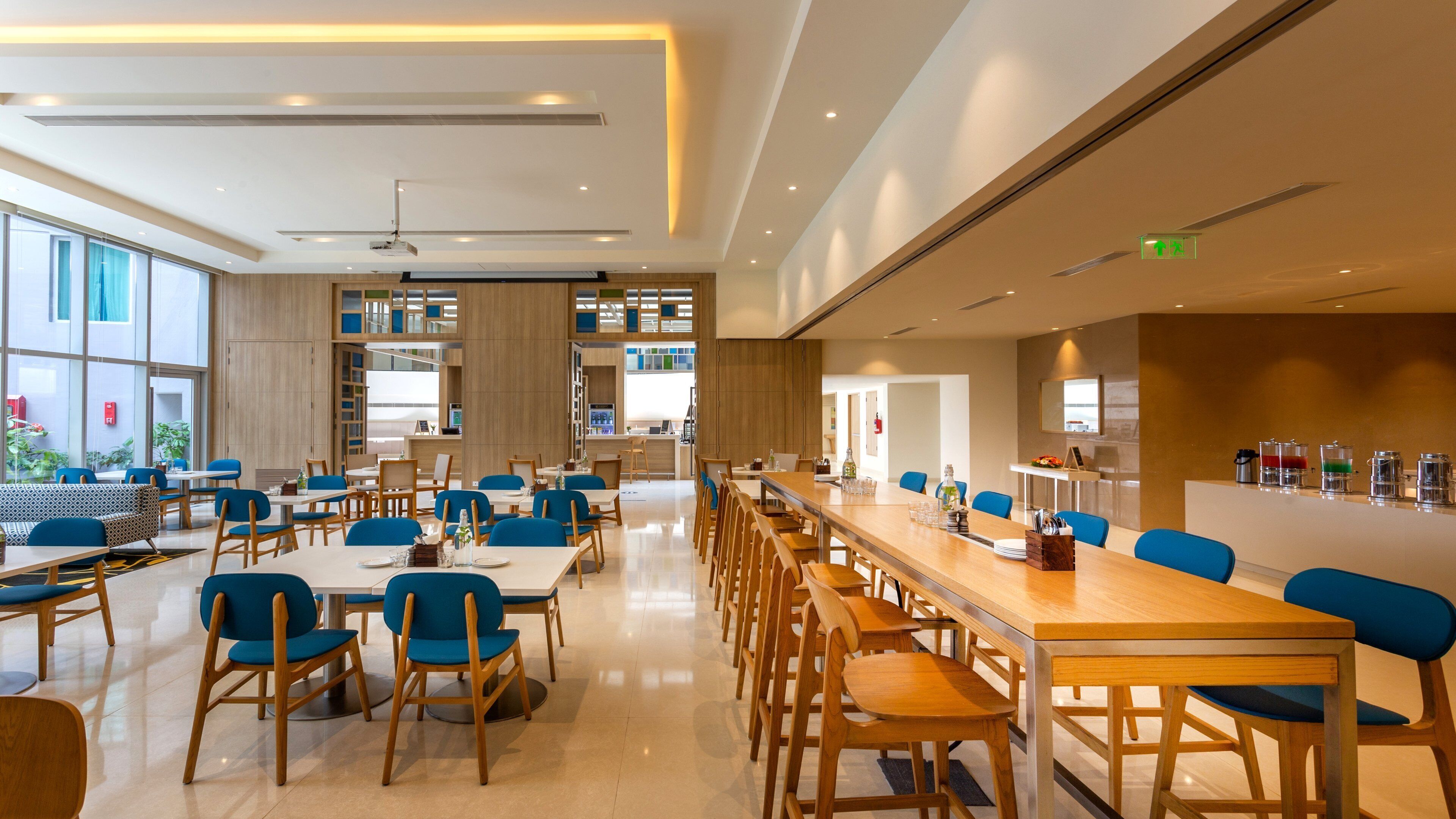 Un vaste espace repas avec de grandes tables et des chaises bleues, un coin cuisine avec un buffet, et une grande fenêtre laissant entrer la lumière naturelle.