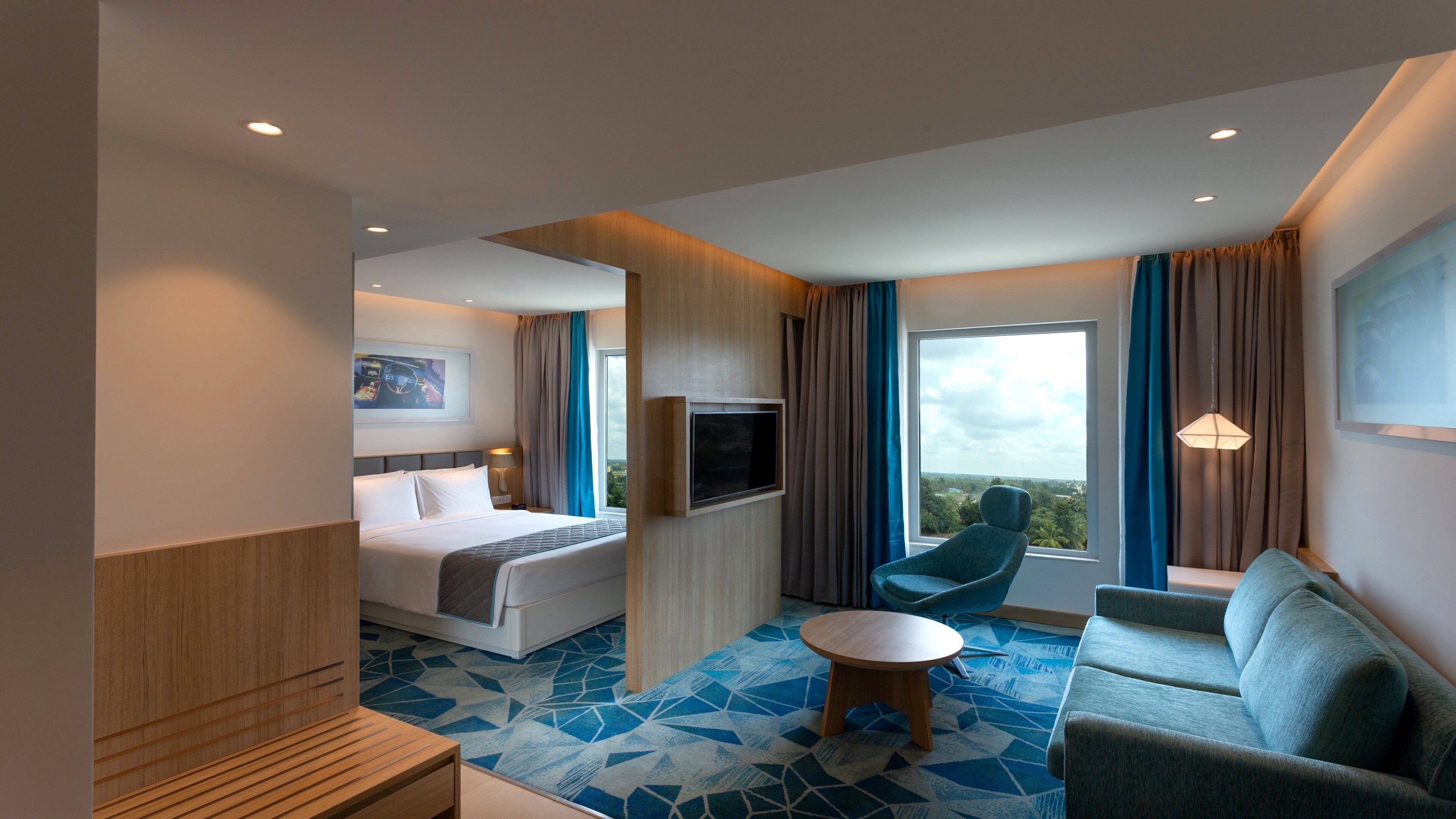 A hotel room with a bed, a TV mounted on the wall, a sofa, a small round table, and a view of the outdoors through the window.
