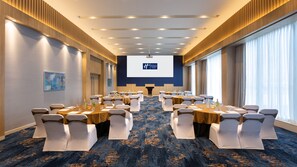 A conference room with rows of tables and chairs, a large screen at the front, and a blue carpeted floor. - Holiday Inn Express & Suites Bengaluru Old Madras Road by IHG (Bengaluru)