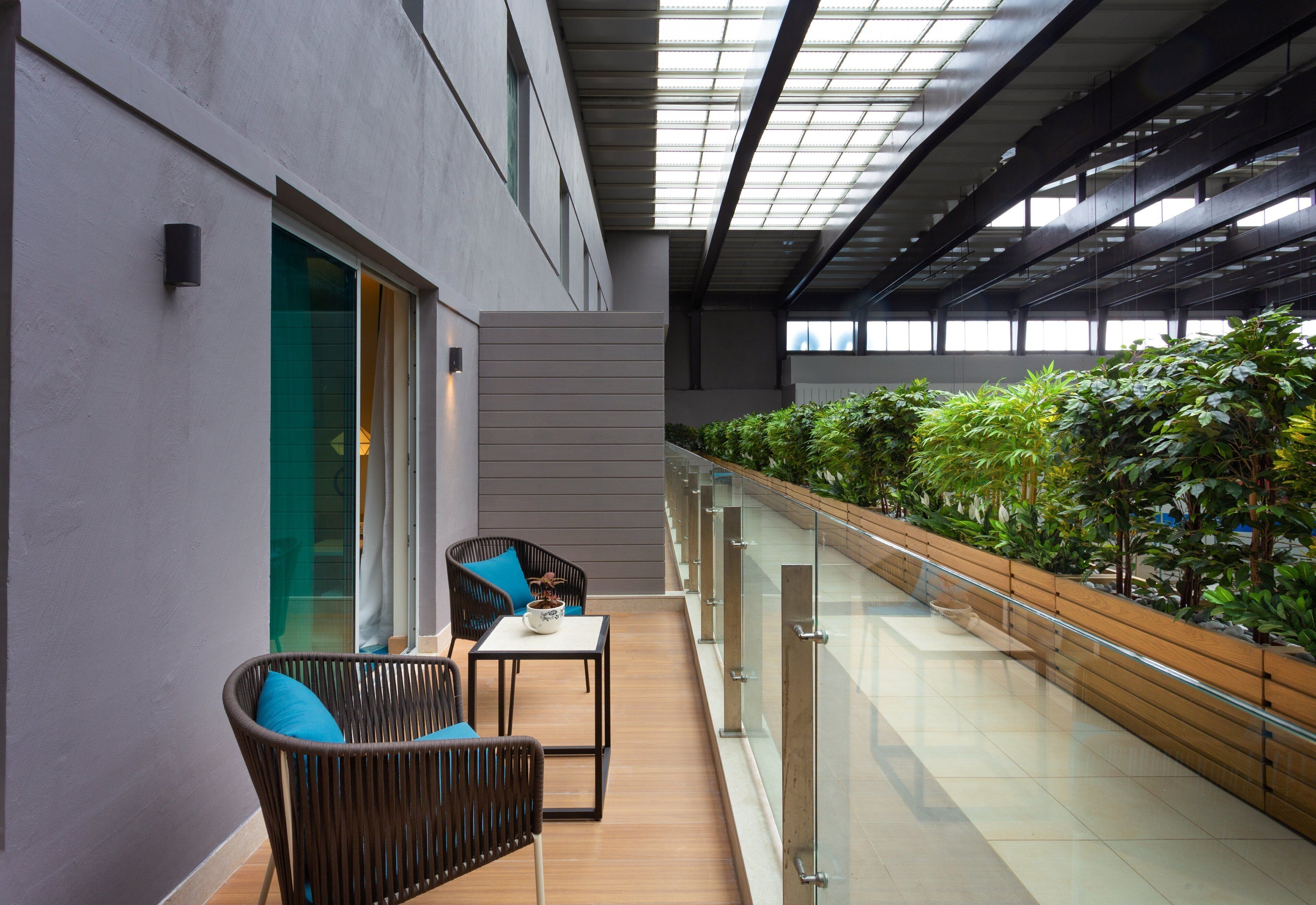Een moderne ruimte met een glazen balustrade, een zithoek met een stoel en tafel, en een rij groene planten langs de balustrade.