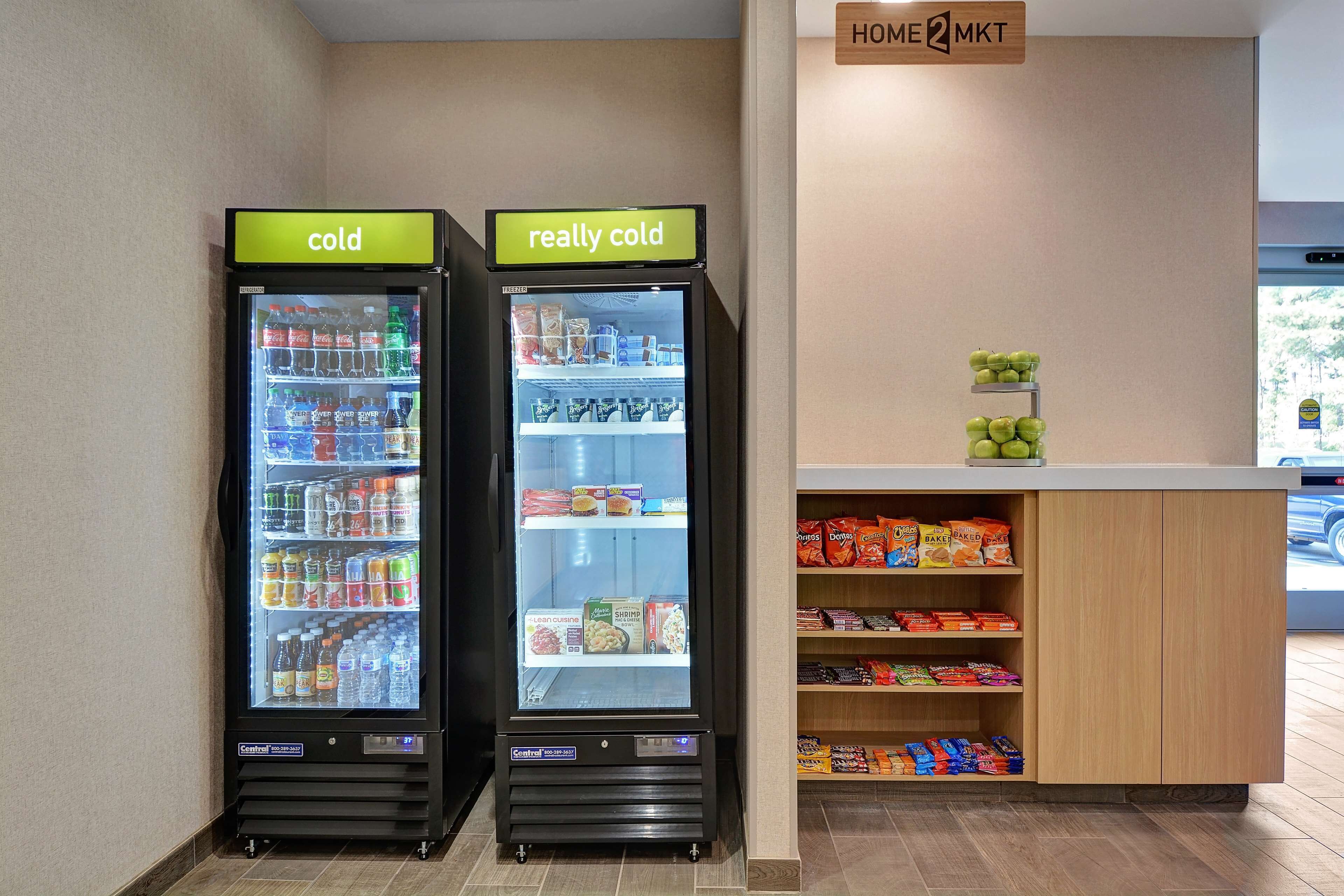 Dans un hôtel, on trouve deux distributeurs de boissons réfrigérés, étiquetés « froid » et « très froid », ainsi qu’un comptoir où sont exposées des collations et une enseigne indiquant « HOME2MKT ».