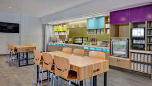 A modern dining area with wooden tables and chairs, a kitchen counter with food display, and a coffee machine. - Home2 Suites by Hilton Fort Mill, SC (Fort Mill)