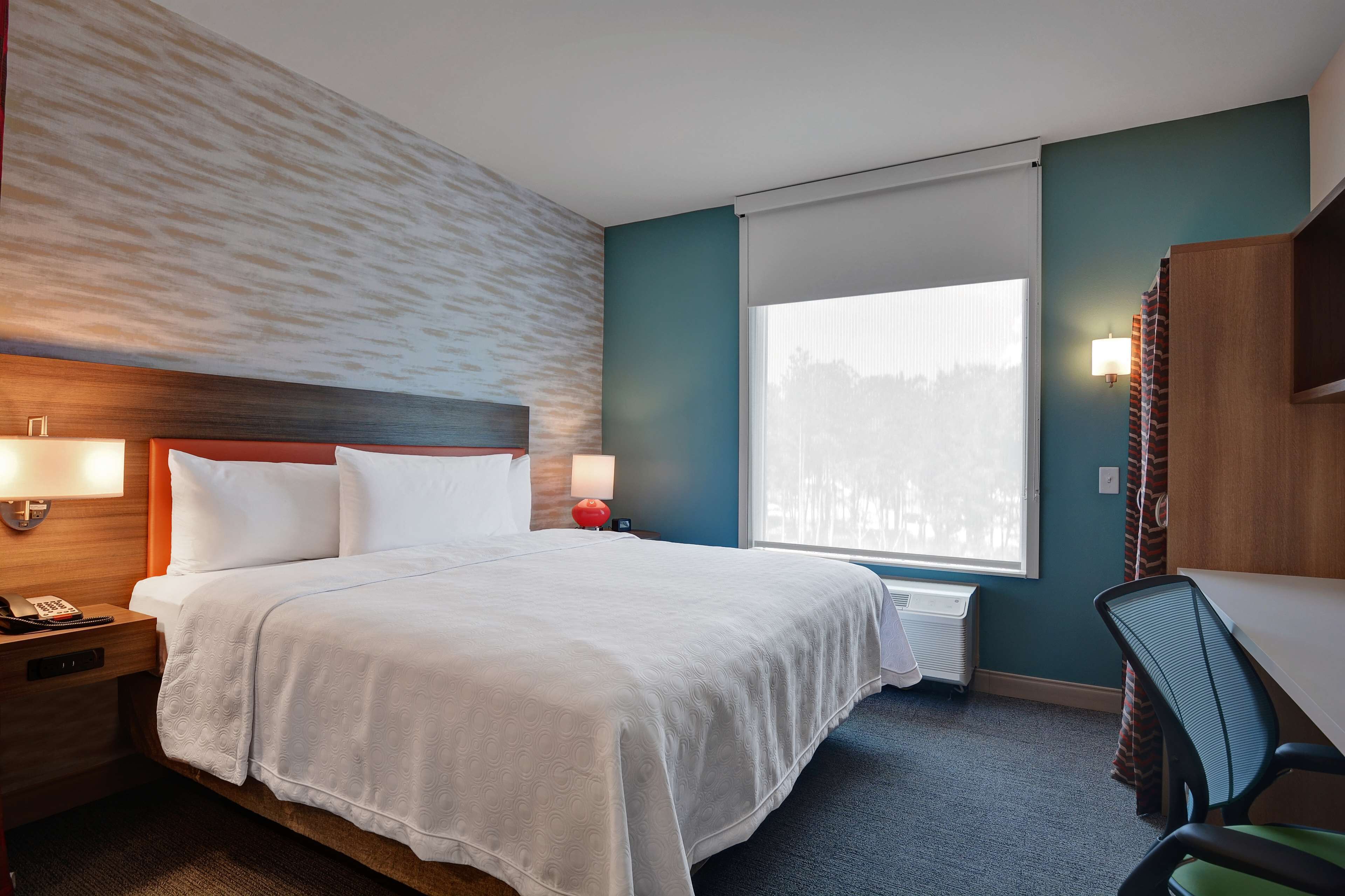 Une chambre d’hôtel comprenant un lit, une table de chevet, une lampe, un bureau, une chaise et une grande fenêtre.
