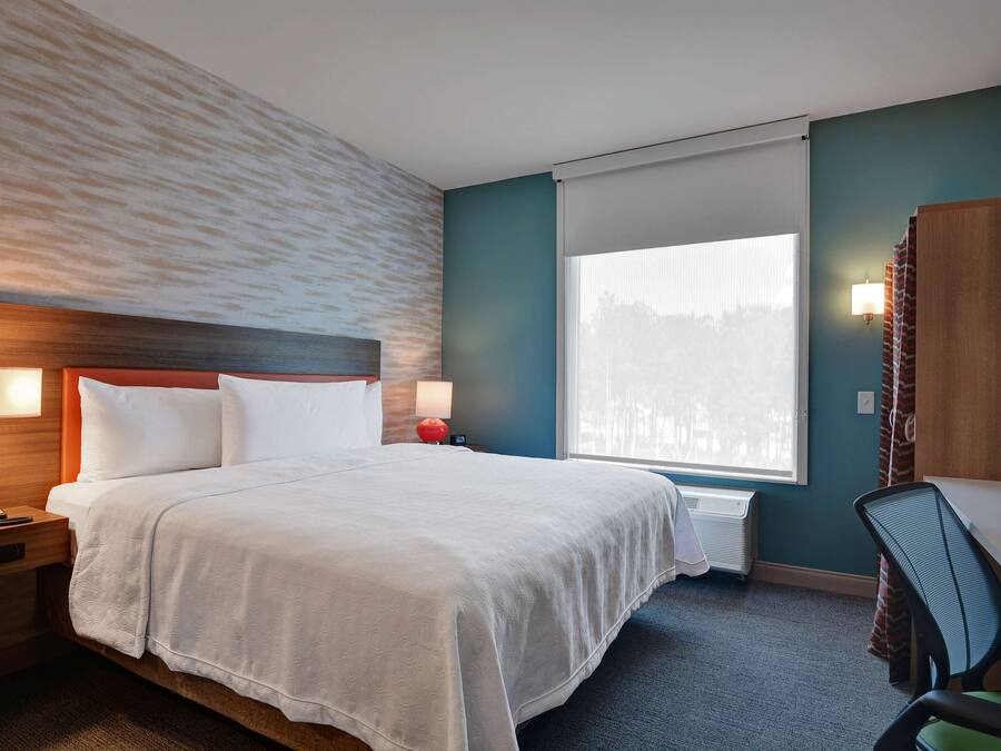 Une chambre d’hôtel comprenant un lit, une table de chevet, une lampe, un bureau, une chaise et une grande fenêtre.