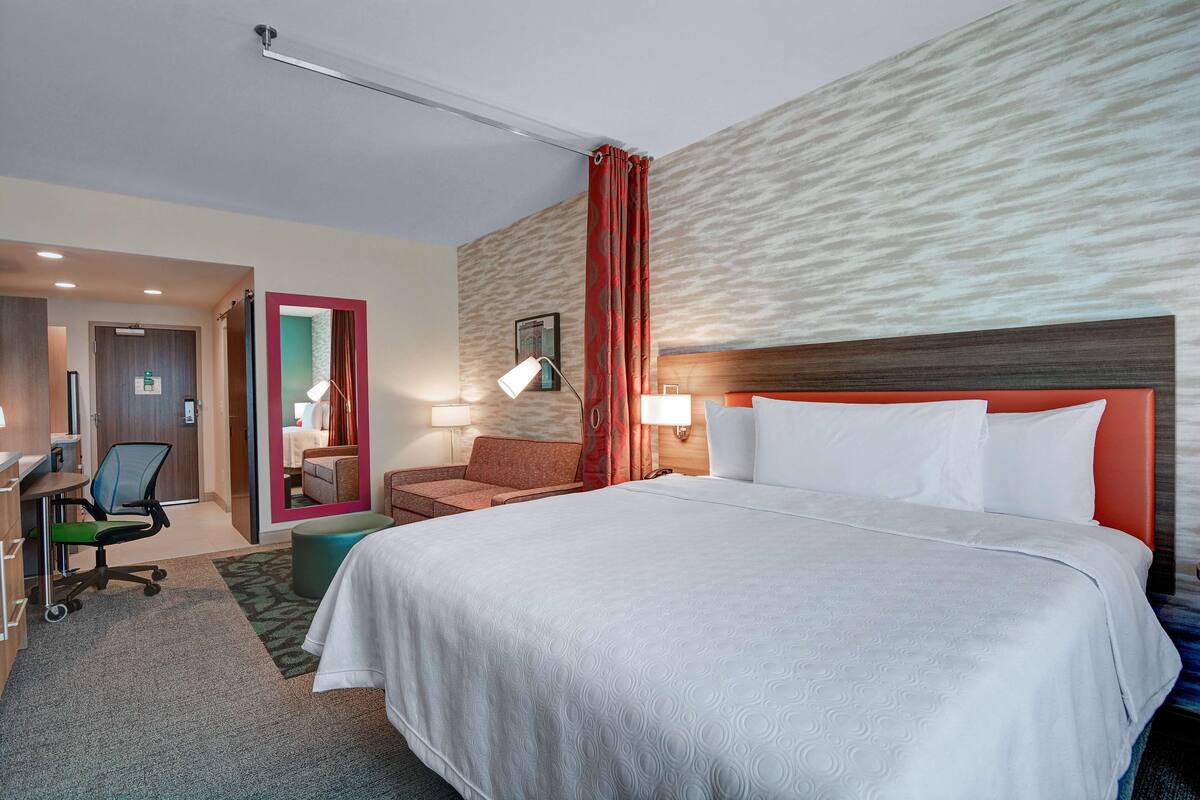 Une chambre d’hôtel avec un grand lit, un bureau avec une chaise, un petit canapé et une salle de bain visible à travers une porte ouverte.