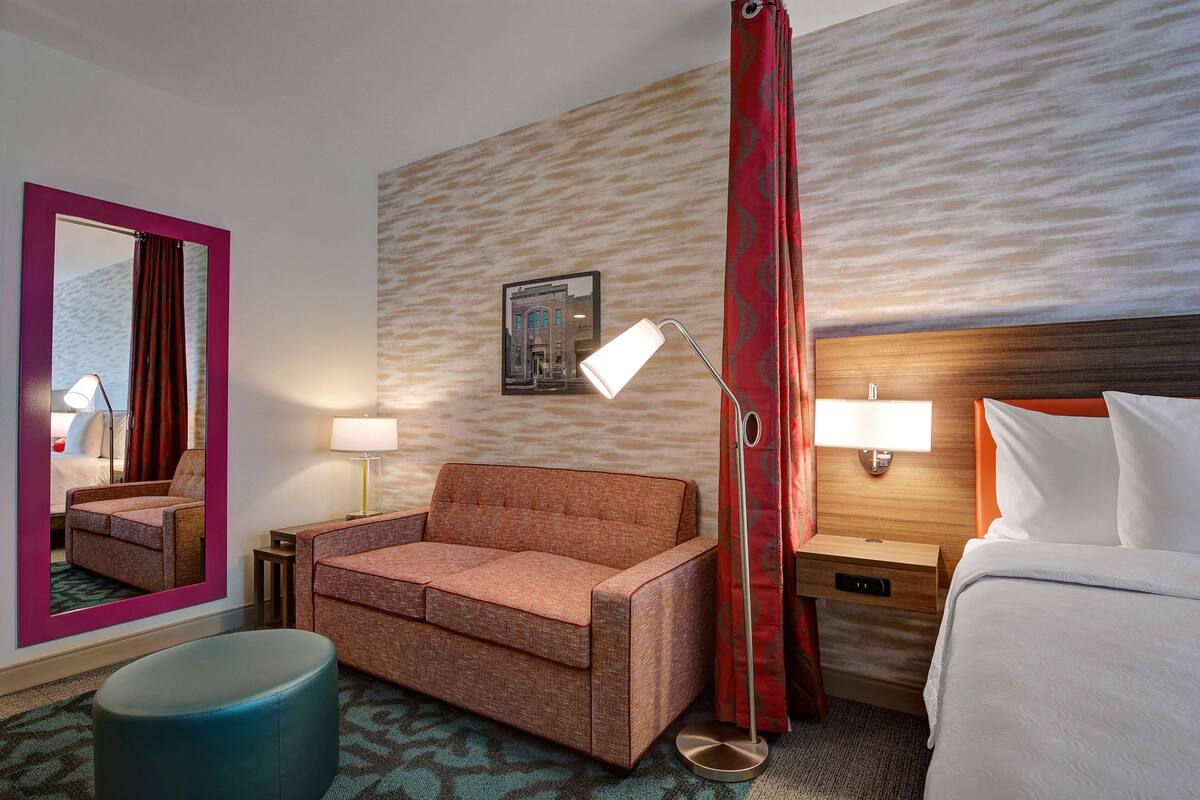 Une chambre d’hôtel avec un canapé, un lit, une table de chevet, une lampe, un miroir et un tableau au mur.